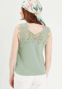 Haut sans manches vert avec un corps texturé et des détails en dentelle florale à l'arrière. Le modèle porte un foulard à motifs qui complète la tenue.
