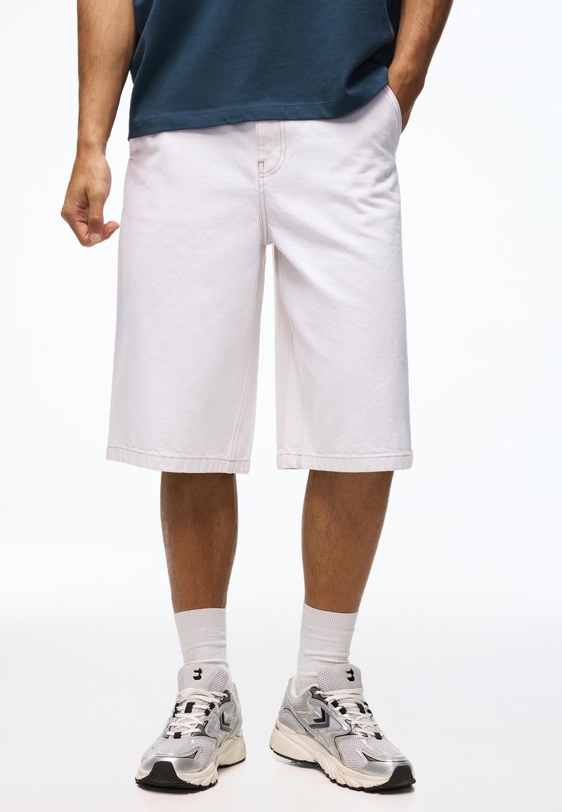 Homme portant un short rose clair jusqu'aux genoux, des chaussettes blanches classiques, des chaussures de course argentées et blanches, et une chemise marine avec une main dans la poche.