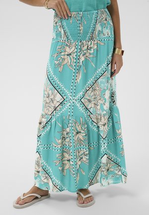 Türkisfarbener Maxirock mit Blumenmuster und gerafftem Bund, getragen mit einem türkisfarbenen Oberteil, goldenen Armbändern und weißen Flip-Flops vor hellem Hintergrund.