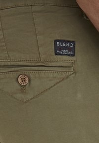 Maslinasto zelene hlače s teksturiranim materijalom, s crnim oznakama na kojima je logo "BLEND" i okruglim uvučenim dugmetom. Uvučen detalj stražnjeg džepa.