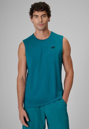 Man with curly dark hair wearing teal sleeveless shirt and matching shorts standing against gray background.