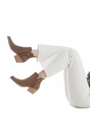 Botines marrones perforados con punta cuadrada, cremallera plateada y tacón de madera apilado, combinados con vaqueros blancos de dobladillo deshilachado.