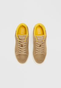 Zapatillas de ante marrón claro con forro de tela amarilla, que presentan cordones beige y una suela de goma blanca. Logo visible en las plantillas.