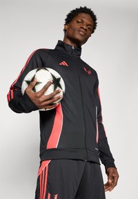 Chaqueta deportiva negra con acentos rojos y ribete blanco; el modelo sostiene un balón de fútbol en blanco y negro. Tejido texturizado para ropa deportiva.