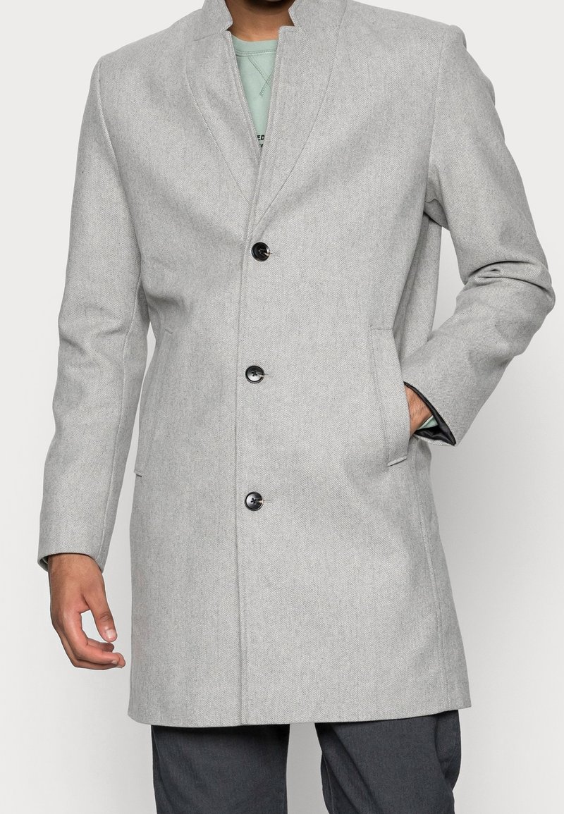 Cappotto a spina di pesce grigio chiaro con rever a contrasto, cinque bottoni neri, tasche laterali e vestibilità strutturata.