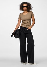 Žena kovrčave kose koja nosi crne sunčane naočale, smeđu majicu kratkih rukava, široke crne hlače i bijele tenisice s platformom, držeći crnu torbicu.