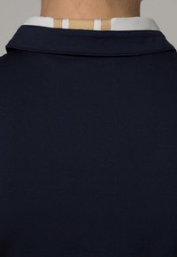 Tummansininen poolopaita, jossa on kontrastina valkoinen kaulus ja kaulan takana beigen sävyinen teksturoitu yksityiskohta. Sileä kangas.