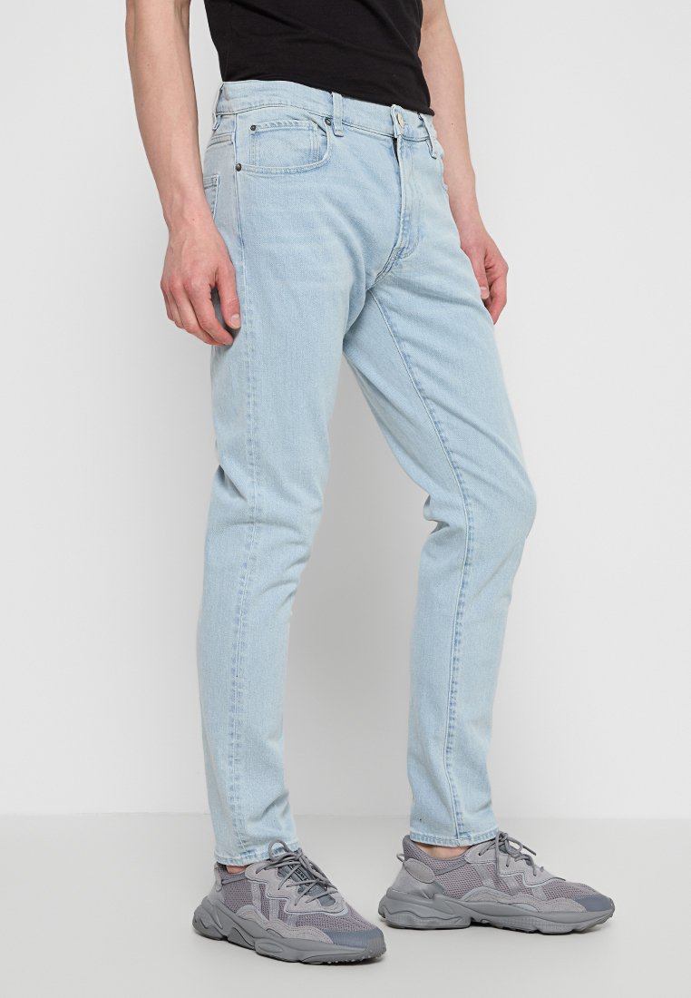 Hellblaue Jeans aus Denim mit einer schmalen Passform, ausgestattet mit klassischem Fünf-Taschen-Stil und einem Knopfverschluss. Getragen mit grauen Sportschuhen.