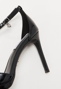 Musta kiiltonahkainen korkokenkä, jossa on hoikka stiletto-korko, nilkkaremmi ja pyöreä varvas. Ominaisuuksiin kuuluu kiiltävä pinta ja vähäiset metalliosat.