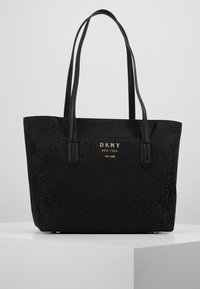 Czarna torba DKNY typu tote z wytłoczonym wzorem logo, gładką fakturą, dwiema długimi rączkami oraz złotą tabliczką z logo z przodu.