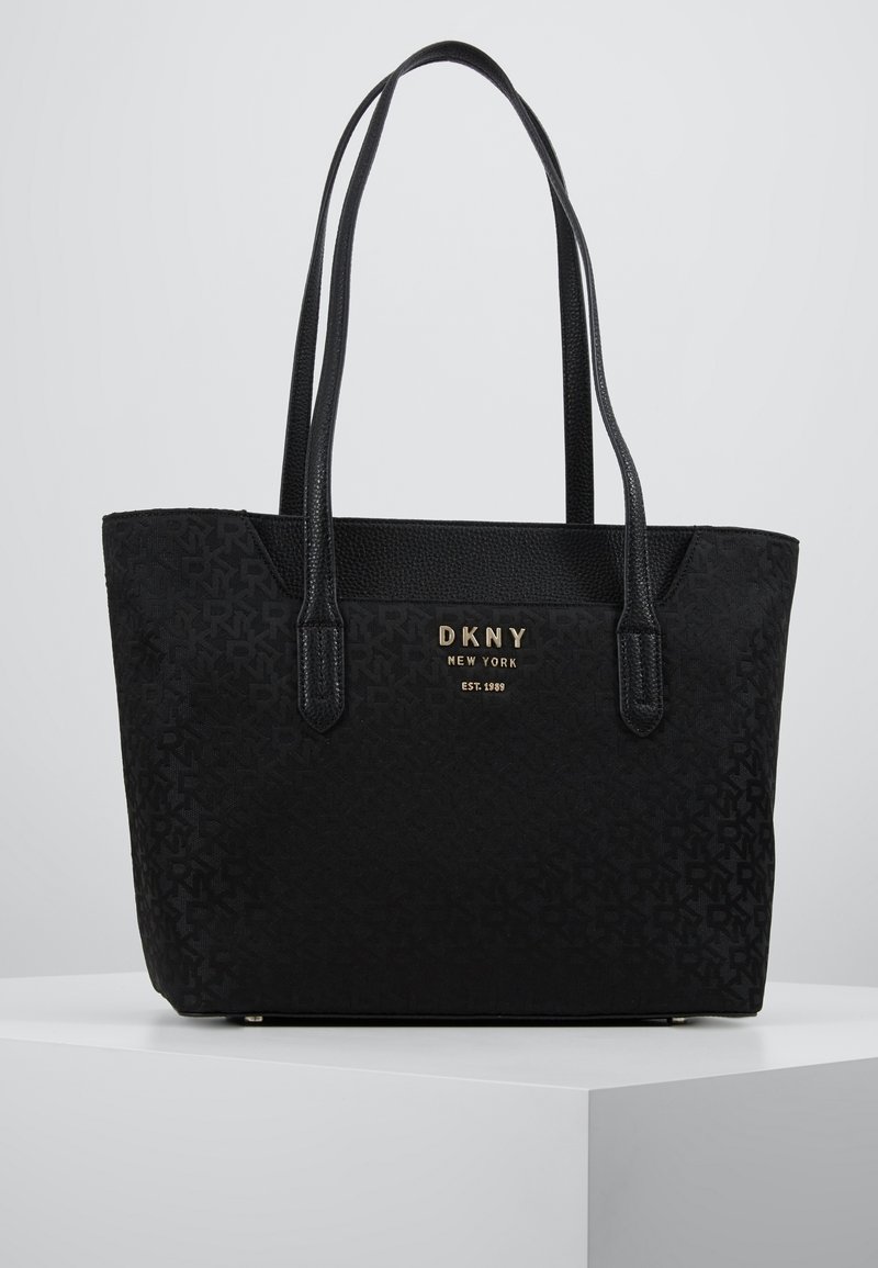 Czarna torba DKNY typu tote z wytłoczonym wzorem logo, gładką fakturą, dwiema długimi rączkami oraz złotą tabliczką z logo z przodu.