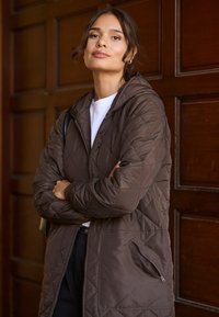 Chaqueta acolchada marrón con capucha, que cuenta con un bolsillo lateral con cremallera. Textura suave y lisa sobre una blusa blanca. Iluminada con luz cálida.