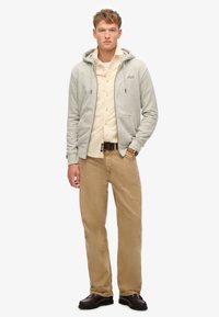 Grå zip-up hoodie med snoretræk, beige skjorte med knapper under, khakisbukser og brune sko. Enkel, afslappet lag-på-lag.