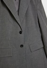Siva blazer s črtami z dvema črnima gumboma, zareznimi ovratniki in žepoma s poklopcem. Narejen je iz teksturirane tkanine za prilagojen videz.