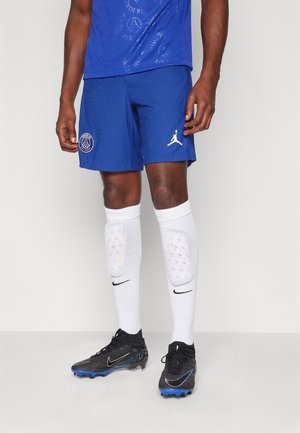 Homme portant un short de football bleu Paris Saint-Germain, des protège-tibias Nike blancs et des crampons Nike noirs avec des semelles bleues sur un fond uni.
