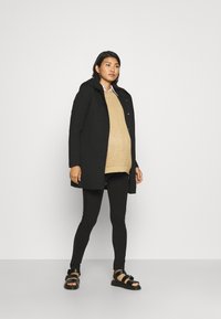 Long manteau noir avec capuche, porté sur un pull en laine beige, des leggings noirs et des sandales noires. Design simple, texture lisse et style minimaliste.