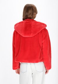 Persona che indossa una giacca con cappuccio finta pelliccia rossa brillante e jeans azzurri, vista da dietro su uno sfondo bianco semplice.