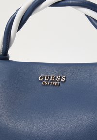 Marineblauwe neplederen handtas met witte en marineblauwe gevlochten handvatten. Heeft een zilveren "GUESS EST 1981" logo plaatje aan de voorkant.