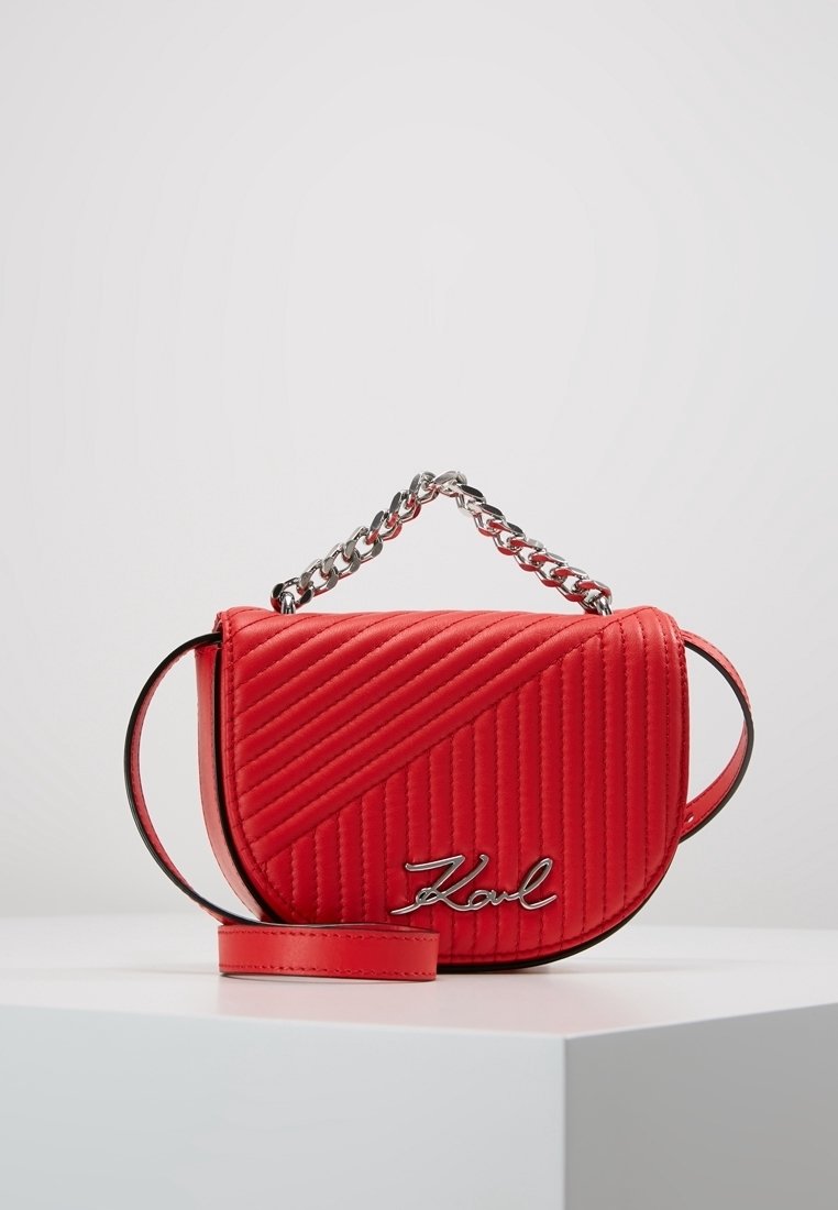 Sac bandoulière matelassé rouge avec poignée chaîne argentée, rabat incurvé et logo « Karl » en argent sur le devant, posé sur une surface blanche.