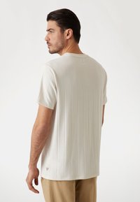Helles beiges T-Shirt mit kurzen Ärmeln, vertikal gestreifter Textur, Rundhalsausschnitt und einem kleinen Logotag an der Unterseite. Weicher Stoff.