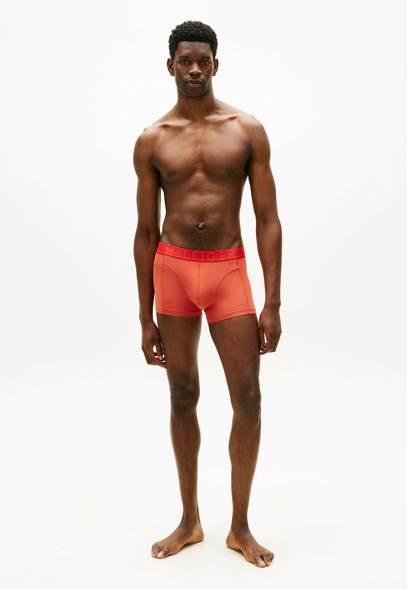 Jeune homme debout à la peau foncée, portant un boxer orange avec la ceinture portant l'inscription « Hilfiger », pieds nus, sur un fond clair uni.