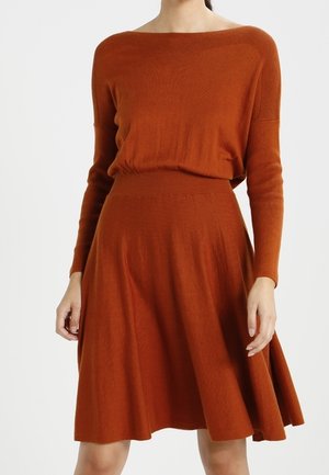 Vrouw draagt een roestkleurige gebreide jurk met een aangesloten taille, lange mouwen en een uitlopende rok, staand tegen een effen witte achtergrond.