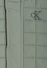 Veste matelassée en vert clair, avec des panneaux cousus et un logo CK brodé en noir sur la poitrine. Texture lisse.