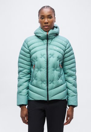 Femme portant une veste matelassée bleu sarcelle avec capuche, poches zippées et pantalon noir, debout devant un fond blanc uni.