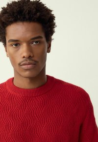 Piros kötött pulóver hullámmintával, bordázott kerek nyakkivágással és texturált anyaggal. A modell rövid, göndör hajjal rendelkezik.