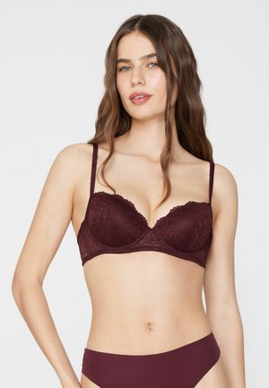 Mujer que lleva un juego de sujetador de encaje burdeos oscuro a juego y ropa interior lisa, con cabello largo y ondulado de color marrón, sobre un fondo liso.