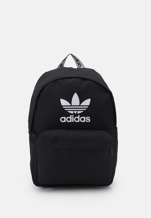 Mochila negra de Adidas con bolsillo delantero con cremallera, logo de trébol y asa superior con marca, mostrada sobre un fondo blanco.