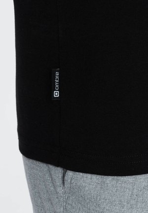Camiseta de algodón negra con dobladillo curvado, que presenta una pequeña etiqueta blanca "ombre" en el costado. Textura suave y diseño ajustado.
