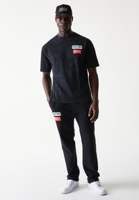 Μαύρο βαμβακερό σετ με μπλούζα και παντελόνι με γραφικά μπαλώματα, υφή υφάσματος και λευκά αθλητικά παπούτσια. Το καπέλο έχει κέντημα λογοτύπου σε μαύρο και κόκκινο.