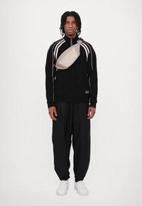 Svart hettejakke med glidelås og hvite striper, løse svarte bukser, og en lys grå crossover-veske, kombinert med hvite joggesko.