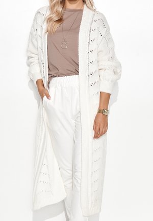 Femme portant un long cardigan blanc en maille par-dessus un t-shirt marron et un pantalon blanc à taille élastique, accessoirisée d'un collier pendentif et d'une montre-bracelet en or.