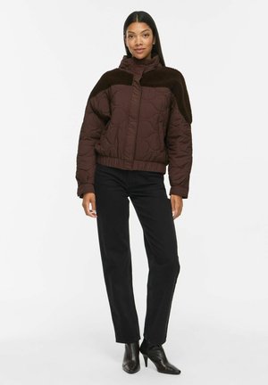Femme debout portant une veste matelassée brun foncé avec un empiècement texturé, un pantalon noir et des bottines noires à talons, sur fond blanc uni.