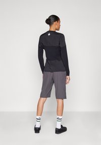Svart langermet topp med en kontrasterende teksturert panel og logo på baksiden, sammen med grå shorts og svarte sko.