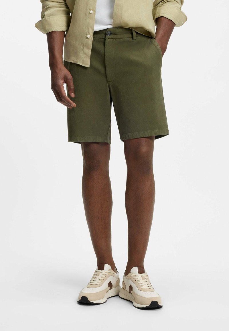Homme portant un short vert olive, une chemise beige boutonnée avec les manches retroussées, des baskets blanches avec des accents marron, debout avec une main dans la poche.