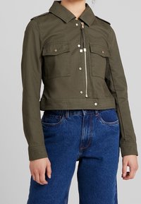 Krótká, ciemnozielona kurtka z bawełny z kołnierzykiem, dwoma kieszeniami na piersi, srebrnym zamkiem błyskawicznym i akcentami guzików. Noszona na niebieskich dżinsach.