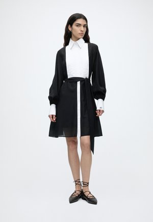 Femme debout portant une robe à manches longues noire et blanche avec un nœud devant, assortie à des ballerines noires à lacets avec des clous.