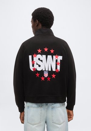 Άτομο που φοράει μαύρο μπουφάν με το «USMNT» και κόκκινα αστέρια γύρω του, συνδυασμένο με γαλάζια τζιν, στραμμένο με την πλάτη σε λευκό φόντο.