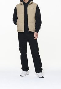 Béžová prošívaná vesta s vysokým límcem, zipem a logem na hrudi, nošená přes černou mikinu s kapucí, doplněná černými cargo kalhotami a bílými teniskami.
