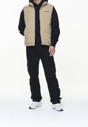 Béžová prošívaná vesta s vysokým límcem, zipem a logem na hrudi, nošená přes černou mikinu s kapucí, doplněná černými cargo kalhotami a bílými teniskami.