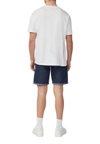 Weißes T-Shirt mit kurzen Ärmeln, dunkle Denim-Shorts mit umgeschlagenen Säumen, weiße Sneakers und Knöchel-Socken, von hinten betrachtet.