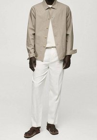 Beżowa kurtka zapinana na guziki, biała koszula, białe spodnie i brązowe sandały. Kurtka ma luźny krój, gładką teksturę i subtelny blask.