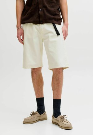 Homme portant un short crème jusqu'aux genoux, des chaussettes noires, des chaussures beige à lacets, et un haut sans manches marron foncé à boutons avec une boucle de ceinture tressée.