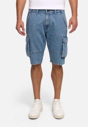 Homme portant un short cargo en denim bleu et des baskets blanches, debout bras détendus le long du corps sur un fond uni.