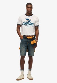Camisa blanca de manga corta con detalles en azul marino y logo, combinada con pantalones cortos de denim, zapatos beiges y un suéter anudado en azul marino y amarillo.