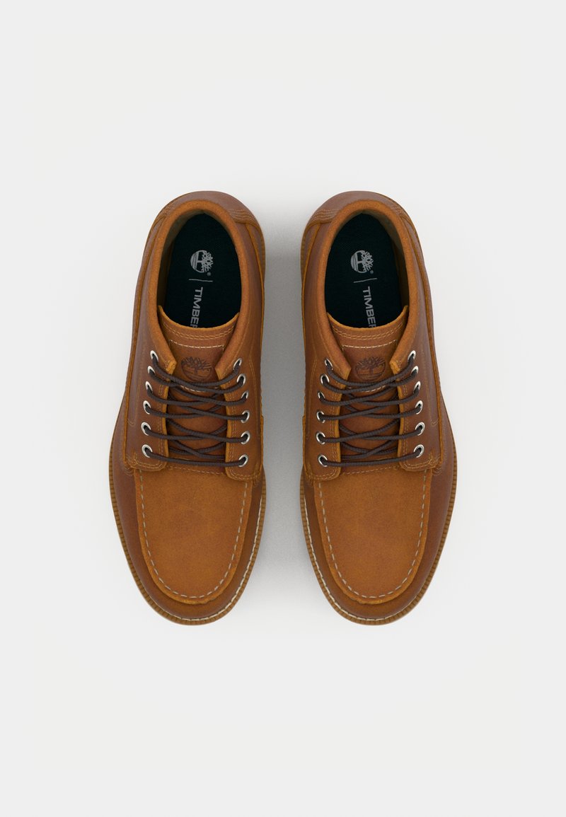 Scarpe in pelle marrone con punta rotonda, cuciture bianche e occhielli in metallo. I lacci sono di un marrone scuro; le solette mostrano il logo.