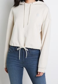 Krämfärgad kort hoodie med dragsko och ribbade ärmslut, med en logodetalj på bröstet och en avslappnad passform. Matchad med blå jeans.